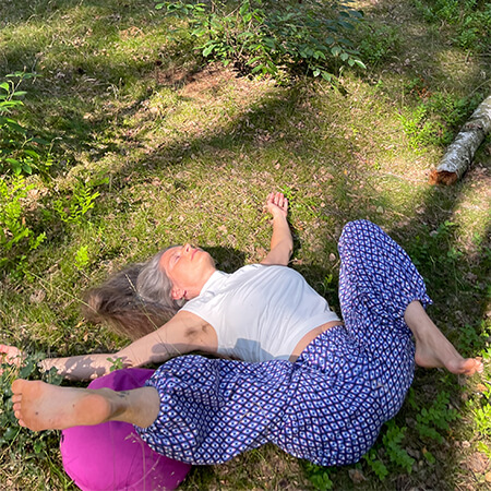 Nirmala lies on her back on the forest floor, one leg bent, the other stretched out to the side, arms open, playful yoga.