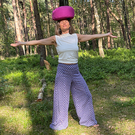 Nirmala stands in the forest, legs wide apart, arms outstretched, pink cushion on her head, practicing mindful, deeply embodied trauma-sensitive yoga.