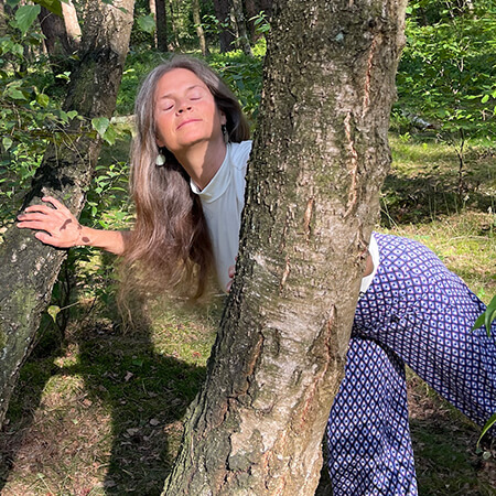 Nirmala looks through two tree trunks, eyes closed, one leg raised, hands on the tree, trauma-sensitive yoga.