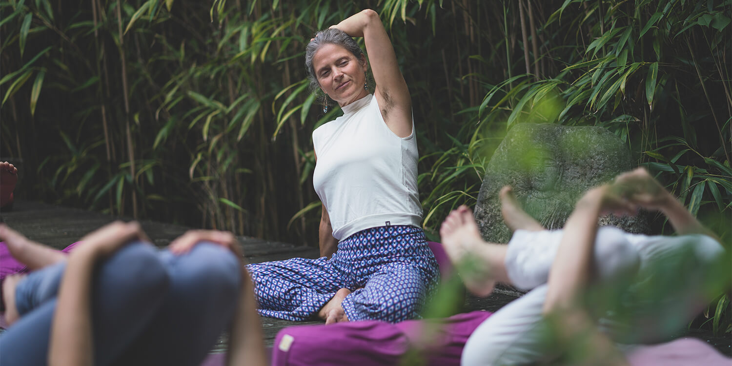 Nirmala unterrichtet traumasensibles Yoga, ein Arm zur Seite gebeugt, draußen vor Bambus, zwei Übende umarmen Knie, zwei pinke Meditationskissen sichtbar.