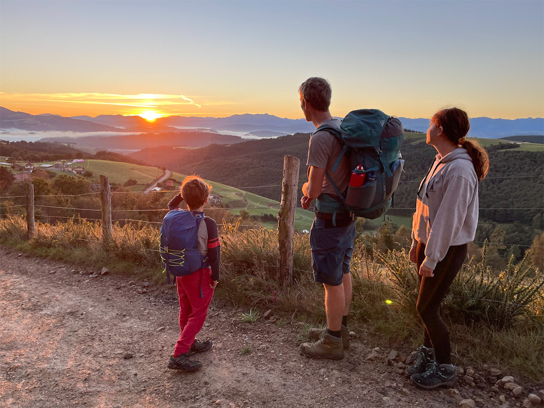 Mann und zwei Kinder mit Rucksäcken betrachten Sonnenaufgang über Bergketten auf Pilgerweg nach Santiago de Compostela.