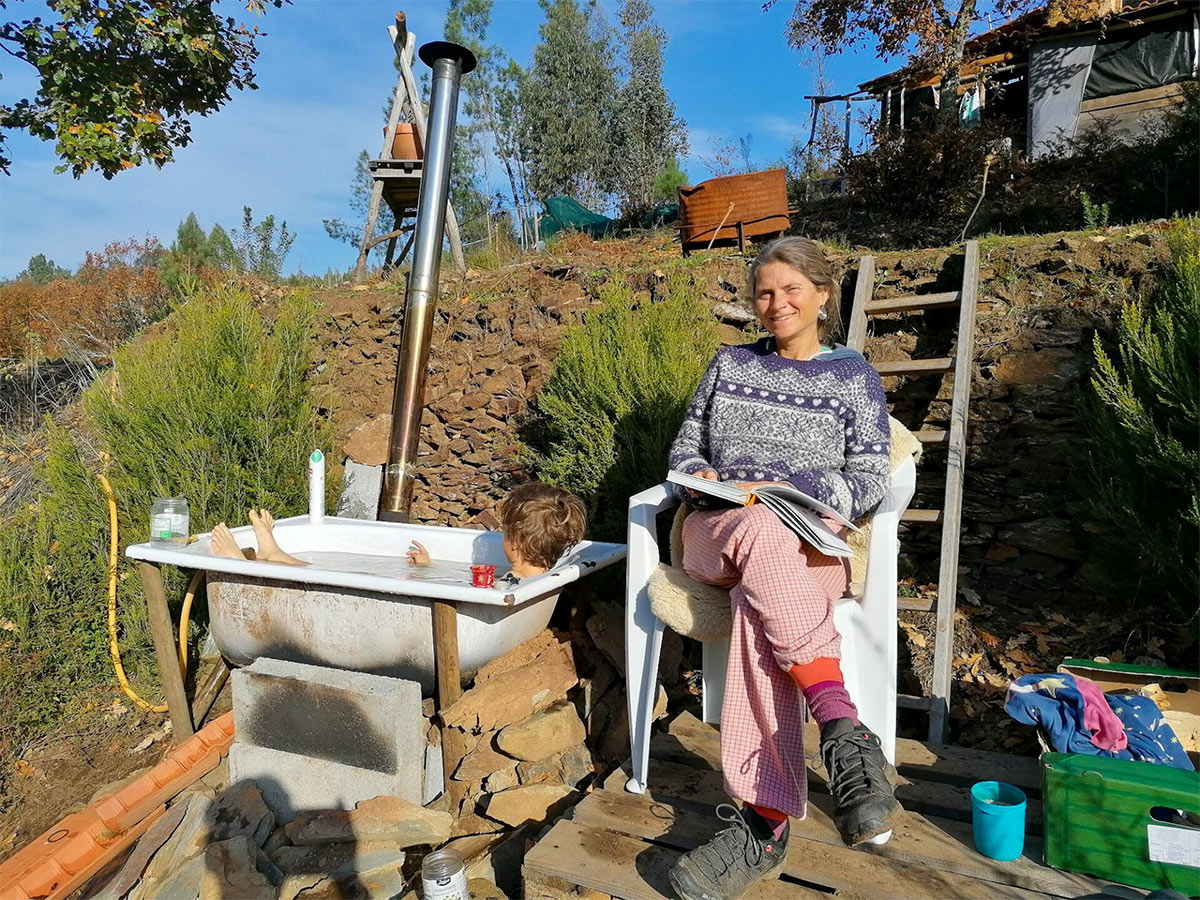 Nirmala sitzt auf einem Stuhl und liest vor, daneben Badewanne mit Person, Ofenrohr führt zum Feuer unter der Wanne, draußen, blauer Himmel.