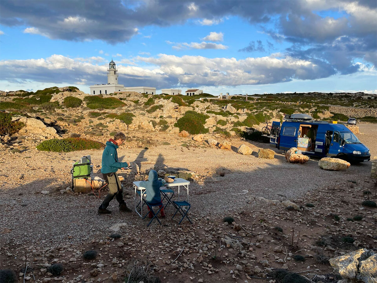Blauer Campingbus in Menorcas felsiger Küstenlandschaft, Lukas und Sohn kochen am Tisch, Leuchtturm im Hintergrund.