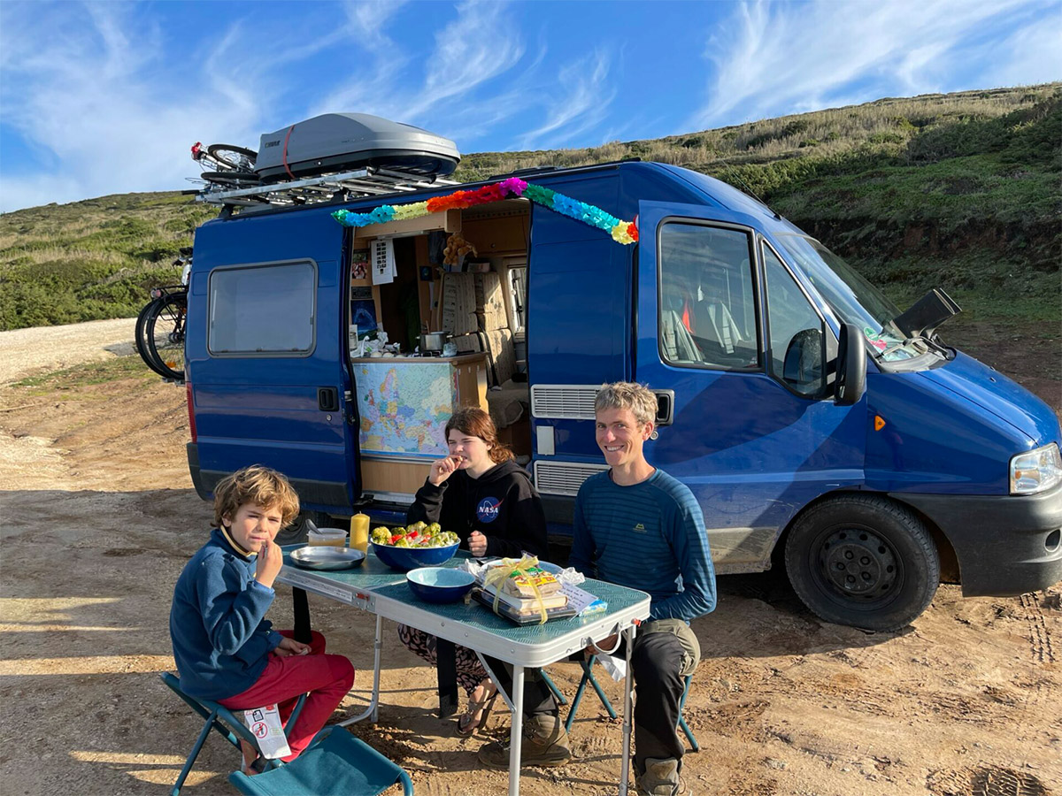 Nirmalas Familie sitzt am Campingtisch vor blauem Bus, dekoriert für Lukas’ Geburtstag, in den Dünen Portugals.