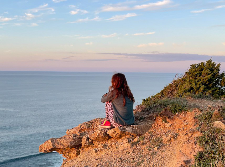 Mädchen sitzt auf Klippe in Portugal, Beine angezogen, betrachtet Sonnenuntergang über dem Meer – Ruhe, Natur, Weite.