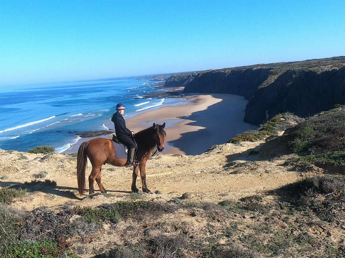Junge Frau auf einem Pferd an der Steilküste Portugals, tiefblaues Meer im Hintergrund