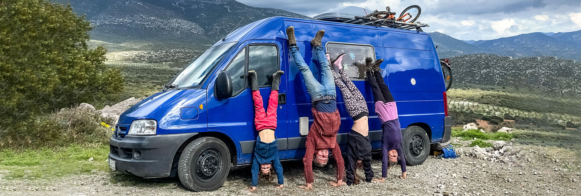 Nirmala, her husband, and two of their children are doing handstands next to each other, leaning against the blue camper van. In nature. 