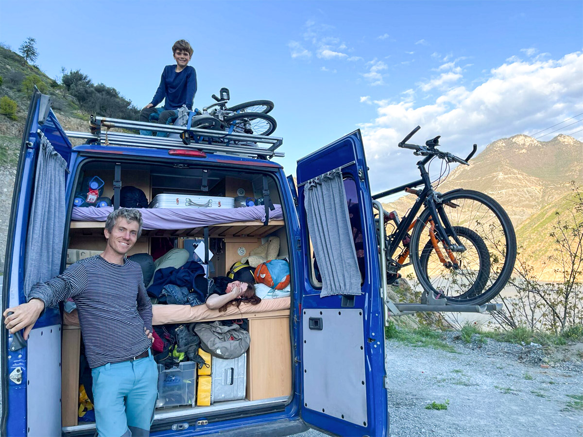 Blue camper van in the mountains, doors open, Lukas leaning against the van, one child sitting inside, the other outside—adventure, nature, family.
