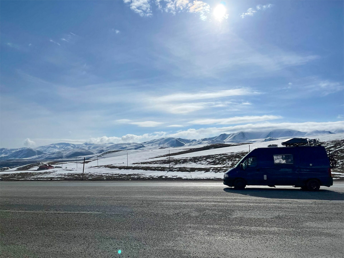 Blue camper van drives along a lonely road through snow-covered mountain landscape – nature, travel, freedom, adventure.