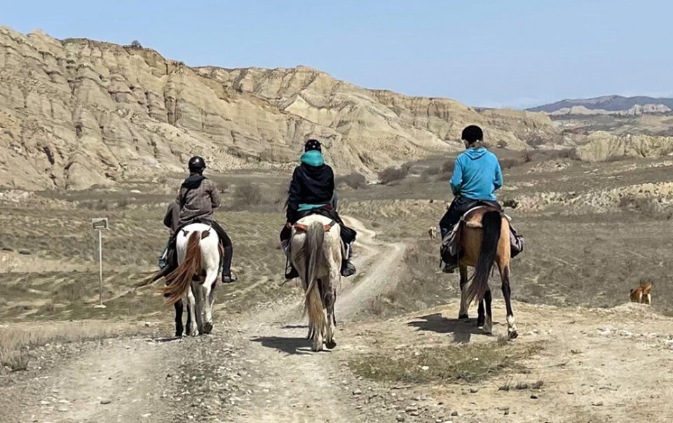 Drei Frauen reiten am Grenzfluss in sandfarbener Steppe und Felsenlandschaft in Georgien, Rückenansicht.