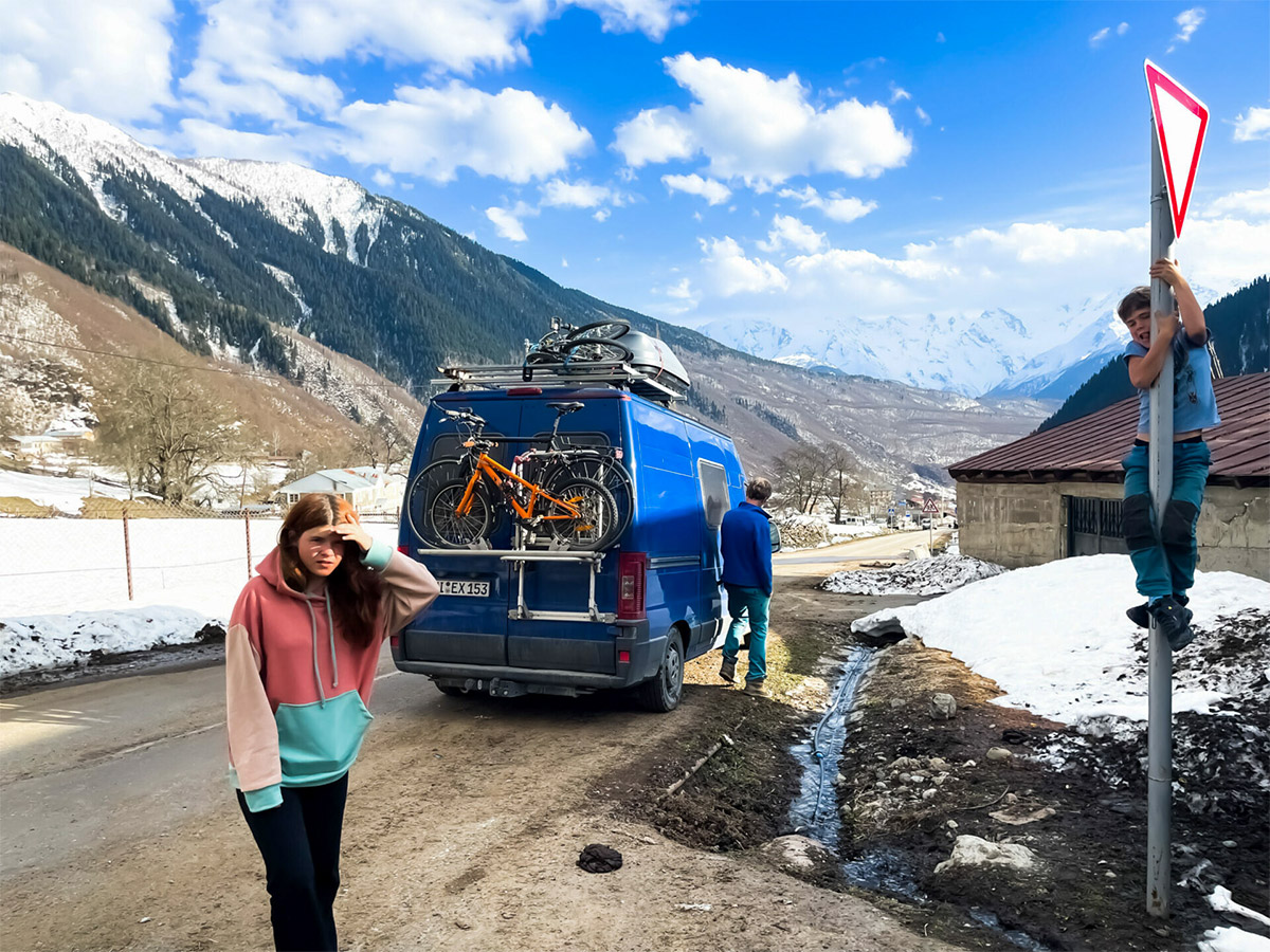 Blauer Campingbus im hohen Kaukasus, verschneite Landschaft, Nirmalas’ Familie zu sehen, Kind klettert an Straßenschild.
