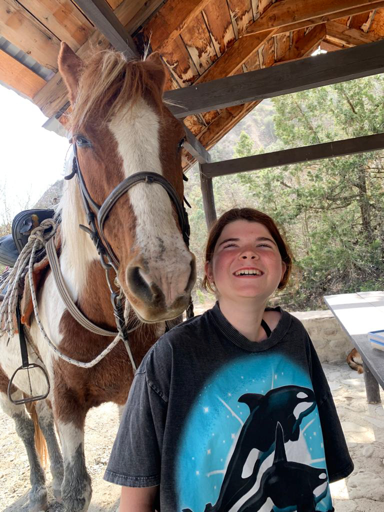 Lachendes Mädchen mit Pferd, dessen Kopf direkt neben ihrem liegt – vertraut, lebendig, naturverbunden.