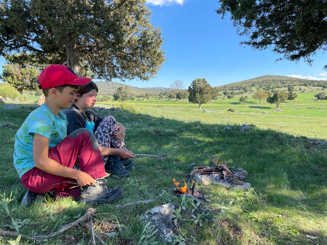 Two children sit next to each other in green, hilly countryside in front of a small campfire on the ground.