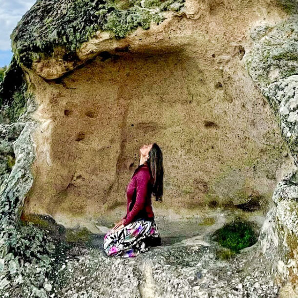 Woman in yoga position Vajrasana on a high rock, looking up into the sky, in the background a green rocky landscape.