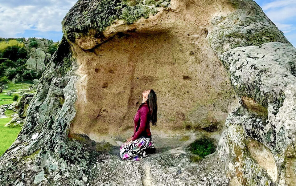 Frau in Yogaposition Vajrasana auf hohem Felsen, Blick nach oben in den Himmel, im Hintergrund eine grüne Felsenlandschaft.