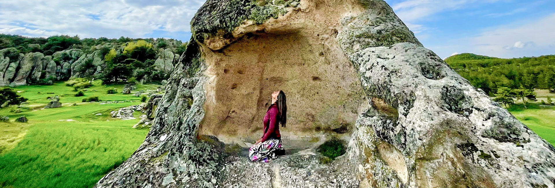 Woman in yoga position Vajrasana on a high rock, looking up into the sky, in the background a green rocky landscape.