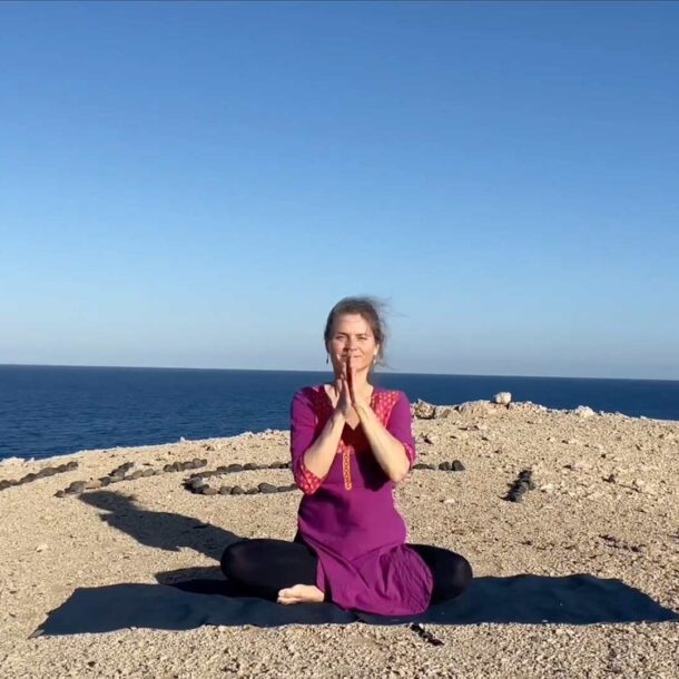 Nirmala sitzt in gekreuzter Position auf Yogamatte, Hände im Anjali-Mudra, auf Sandhügel mit „Jyoti“ aus Steinen, blaues Meer im Hintergrund.