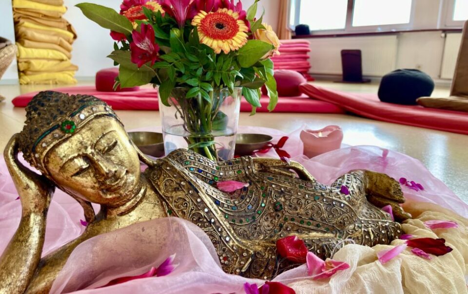Goldene, mit bunten Steinen verzierte Buddha-Statue liegt, Kopf auf Hand gestützt, auf hell-lila Tüchern, umgeben von Blüten, lila Blumen und Kissen im Hintergrund.