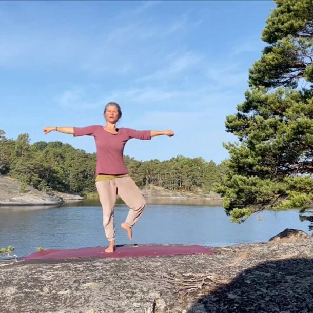 Nirmala steht auf großem Felsen auf roter Yogamatte, ein Bein angewinkelt auf dem Weg zur Baum-Position, Meer im Hintergrund.