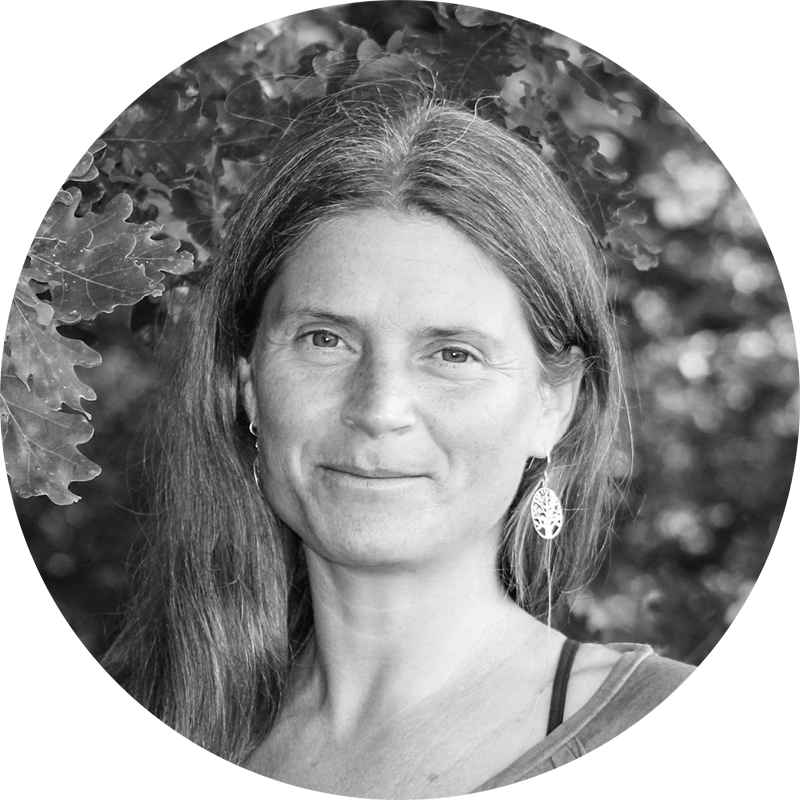 Portrait of Nirmala in black and white, under oak leaves, wearing a filigree tree earring – mindful, meditative, still.