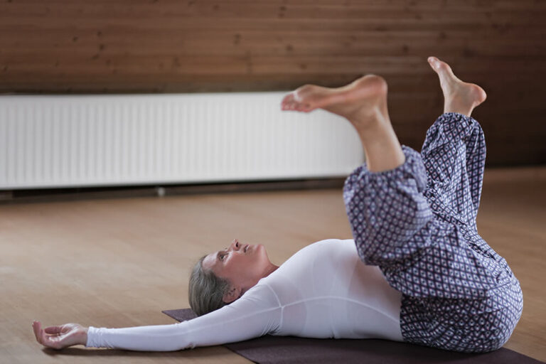 Nirmala liegt quer auf Yogamatte, Arme ausgestreckt, Beine in der Luft geöffnet – traumasensibles, verkörpertes Yoga.