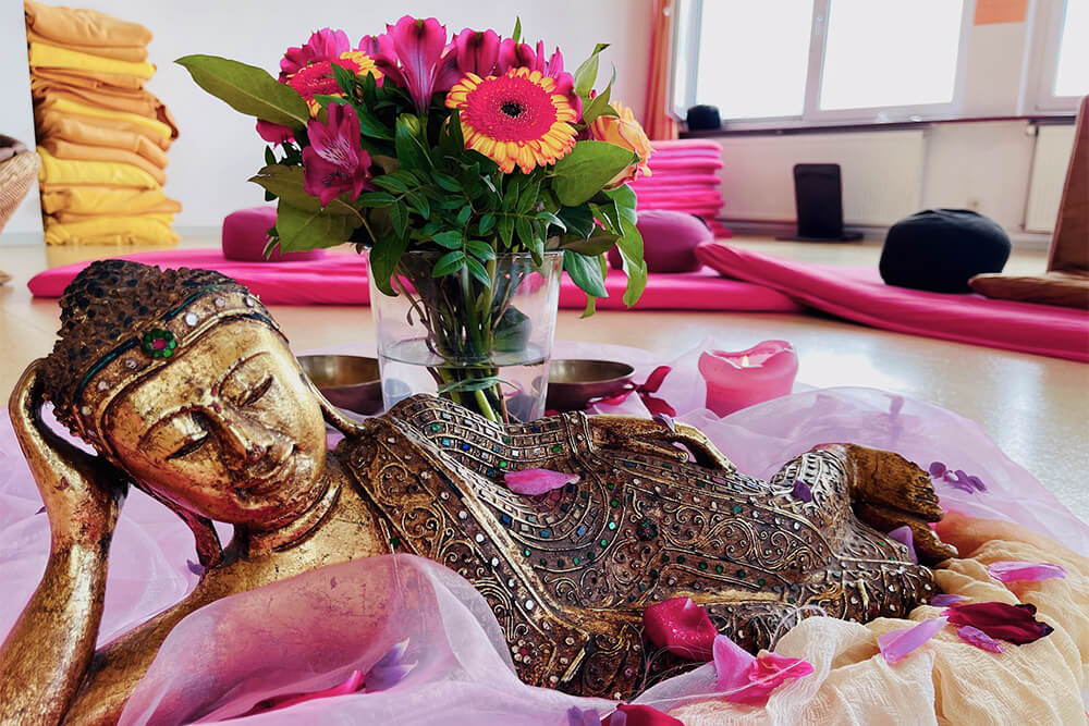 Goldene, mit bunten Steinen verzierte Buddha-Statue liegt, Kopf auf Hand gestützt, auf hell-lila Tüchern, umgeben von Blüten, lila Blumen und Kissen im Hintergrund.