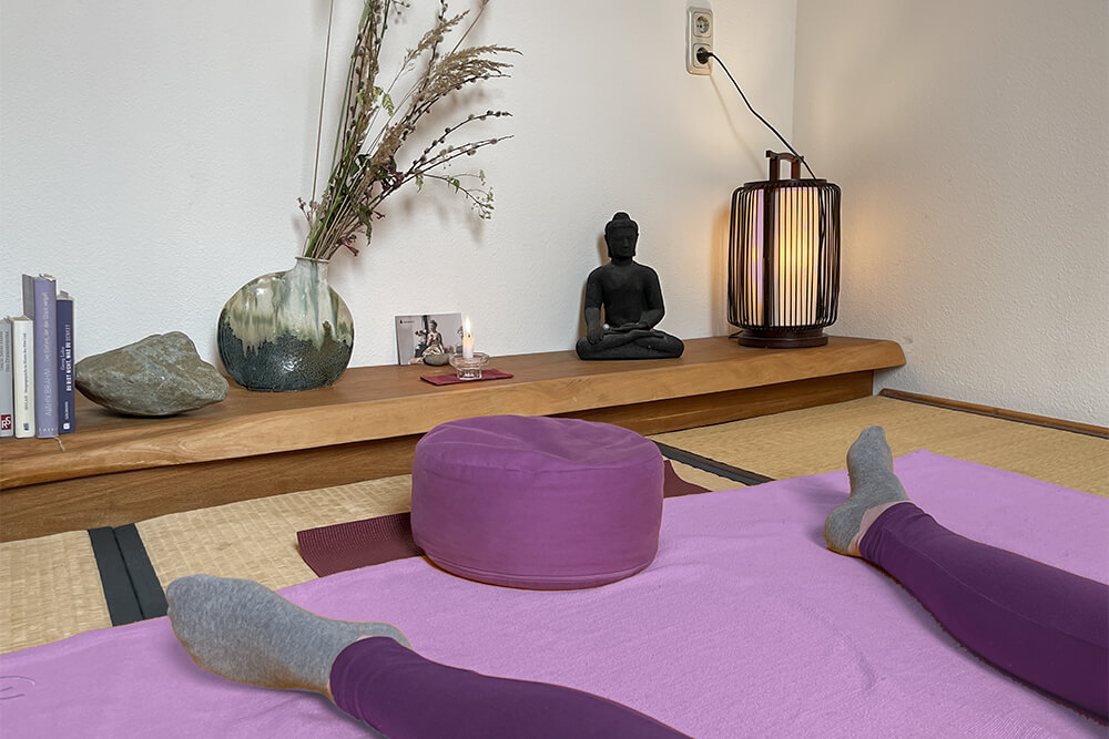 Pinkes Meditationskissen, entspannte Beine auf Decke, im Hintergrund Altar mit Buddha, Büchern und Lampe – Somatic Experiencing, traumasensibel.