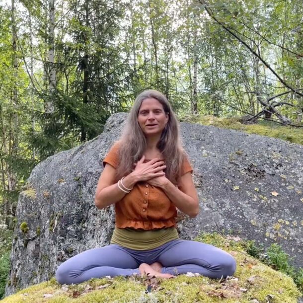 Nirmala sitzt in gekreuzter Meditationshaltung, Hände auf dem Herzen, vor großem Felsen und umgeben von Natur.