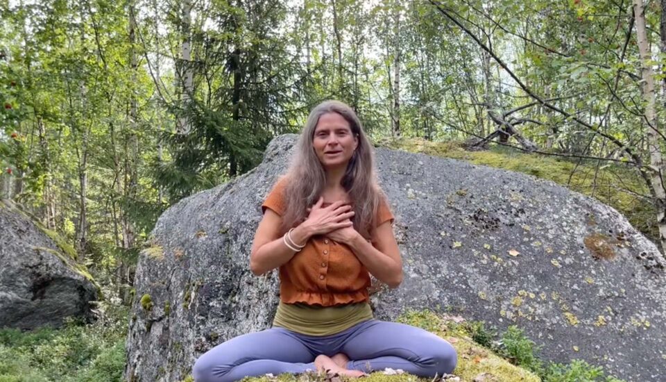 Nirmala sitzt in gekreuzter Meditationshaltung, Hände auf dem Herzen, vor großem Felsen und umgeben von Natur.