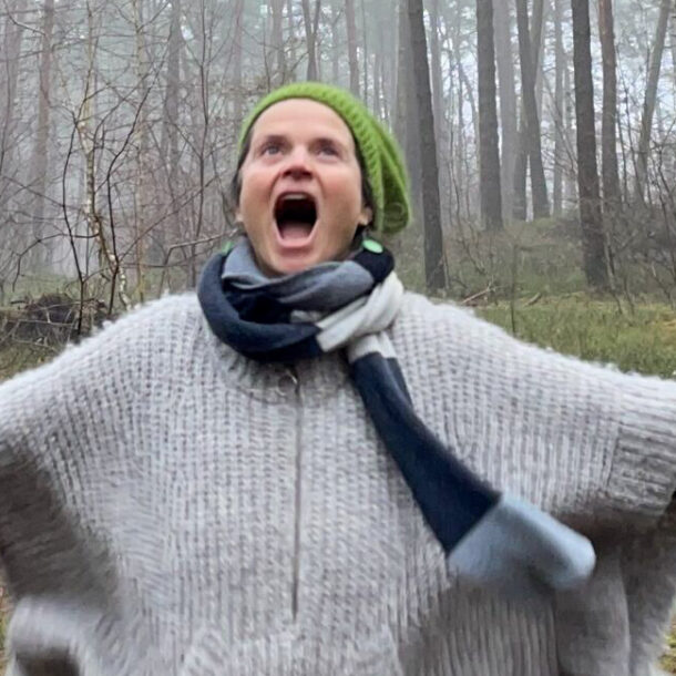 Nirmala runs with outstretched arms through a foggy winter forest, wearing a hat, gloves, and a gray wool poncho, mouth wide open.