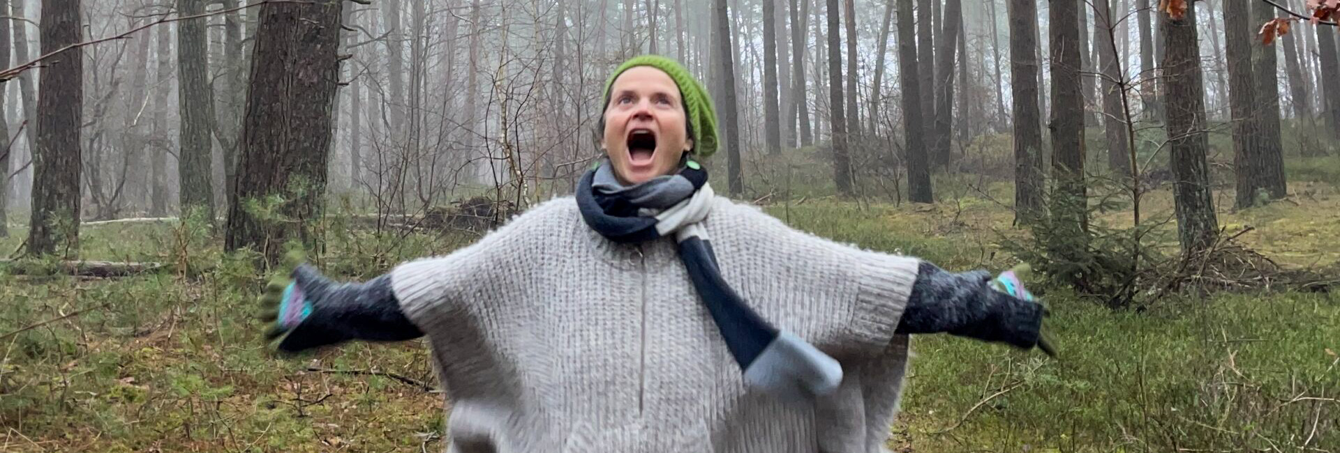 Nirmala rennt mit ausgestreckten Armen durch nebligen Winterwald, trägt Mütze, Handschuhe und grauen Wollponcho, Mund weit geöffnet.