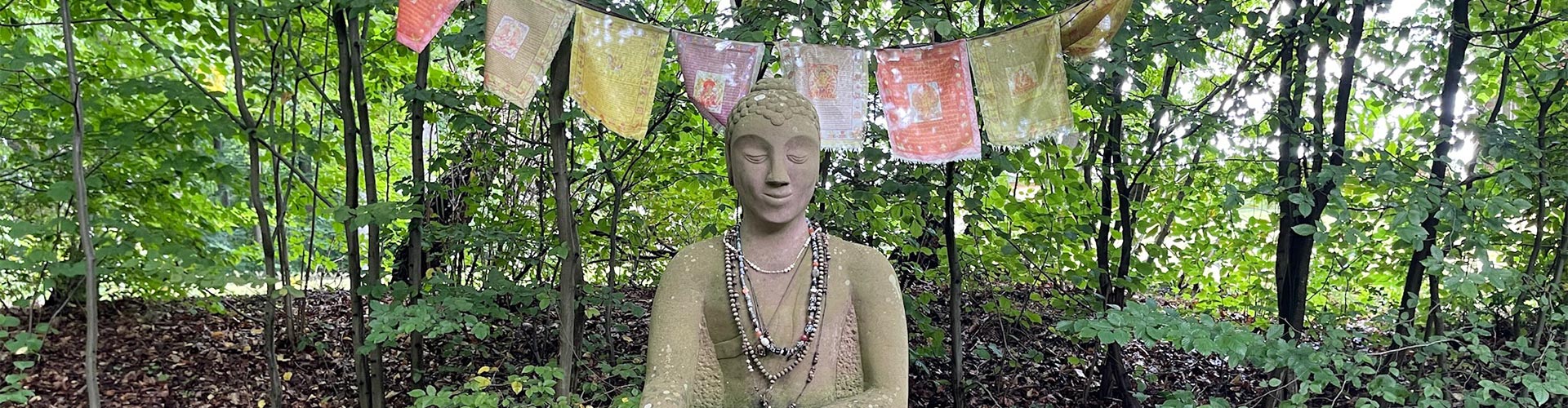 Steinerne Buddhastatue im Park des Haus der Stille, umgeben von Natur, im Hintergrund tibetische Gebetsfahnen.