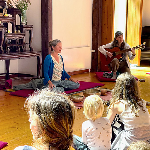 Nirmala unterrichtet im Haus der Stille ein Familienretreat vor Kindern, neben ihr spielt Frau Gitarre, Klangschalen vorne.