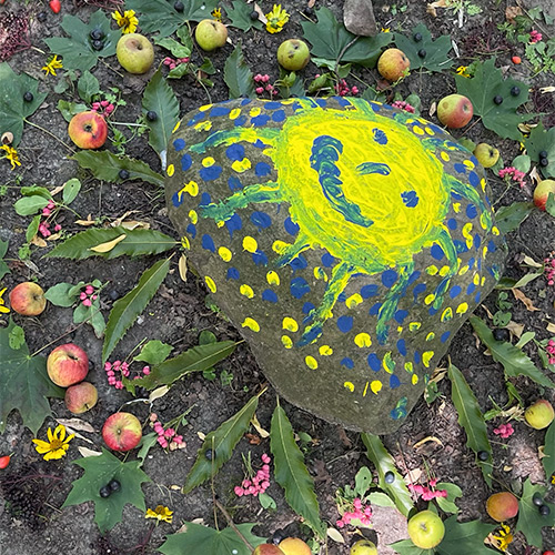 Naturmandala mit großem Stein in der Mitte, darauf lachende Sonne, umgeben von Äpfeln,Blättern, Blüten, achtsam und still.