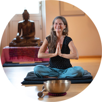 Nirmala sits cross-legged, hands in namaste position in front of her heart, smiling, a large gilded singing bowl in front of her, Buddha in the background.