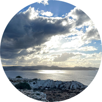 Sunset at the sea, golden rays breaking through the clouds, the sea glistening, gentle mountain ranges in the background.