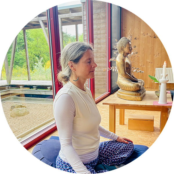 Nirmala in Meditationshaltung vor Altar mit goldener Buddhastatue, große Fenster mit Blick in den Wald im Tempel.