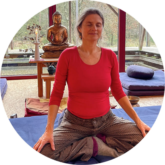 Nirmala meditiert im Tempel vor goldener Buddhafigur auf Altar, verglaste Wand zeigt Wald, meditative Retreat-Atmosphäre.