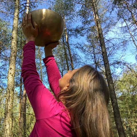 Nirmala seitlich von hinten im Wald, hält große vergoldete Klangschale in die Luft, strahlend blauer Himmel mit Ästen im Vordergrund.
