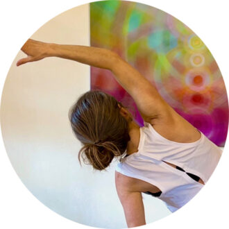 Nirmala from behind in a standing yoga position, one arm bent to the side at her ear, colorful, bright painting in the background.