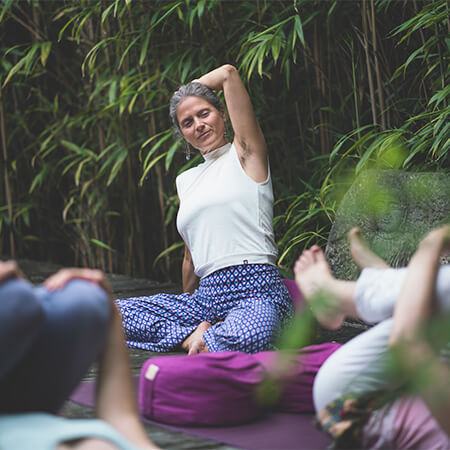 Nirmala unterrichtet traumasensibles Yoga, ein Arm zur Seite gebeugt, draußen vor Bambus, zwei Übende umarmen Knie, zwei pinke Meditationskissen sichtbar.
