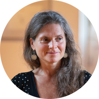Portrait of Nirmala with her hair down, wearing a black dress with white dots and light blue earrings, smiling gently.