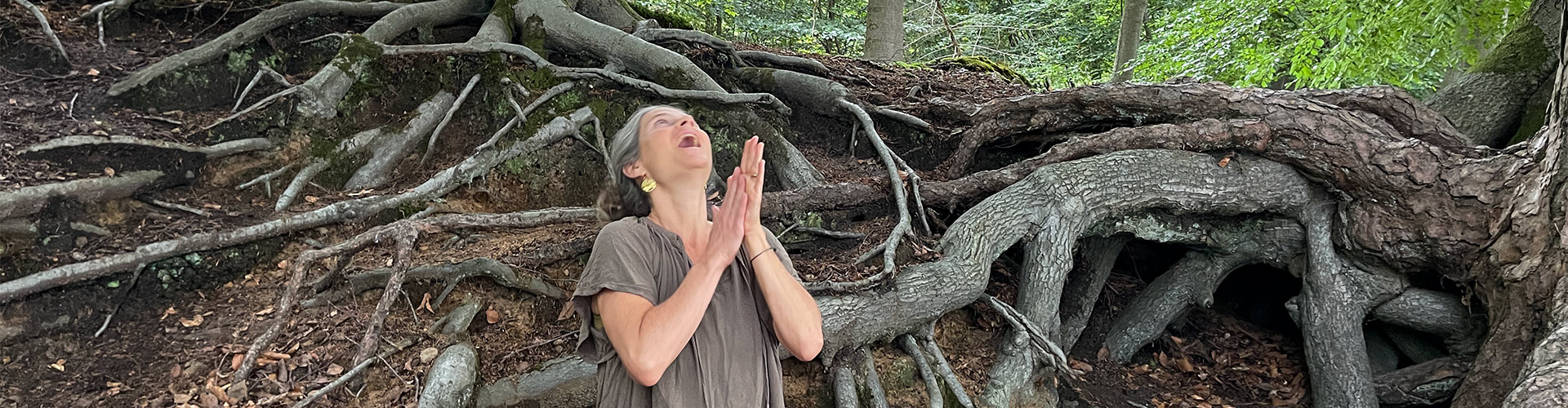 Nirmala sitzt zwischen grossen Wurzeln. Die Hände in Gebetshaltung, lachend – Symbol für Urvertrauen, Geborgenheit, Beckenbodenyoga.