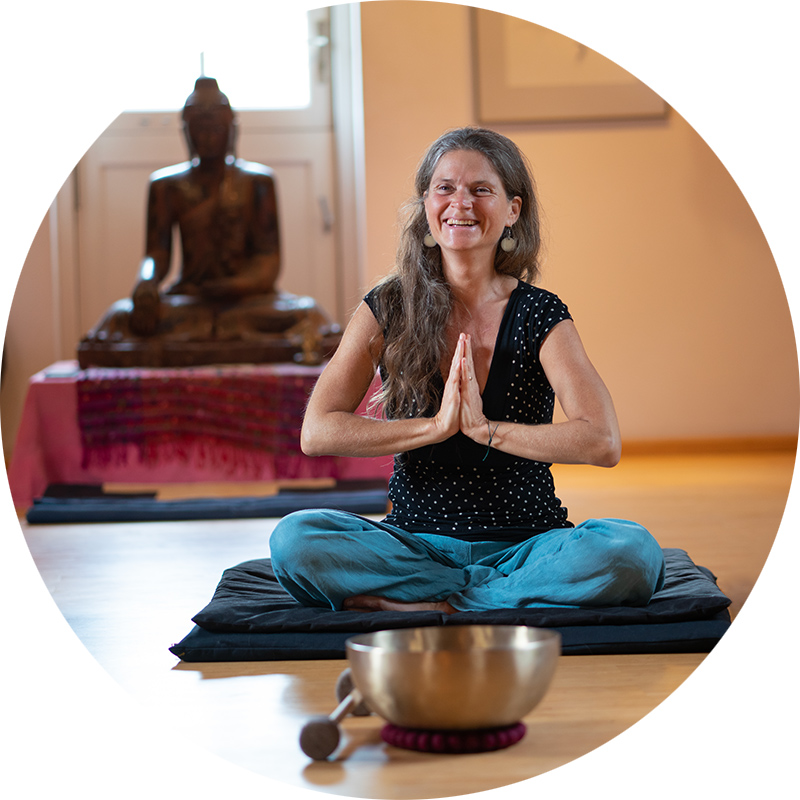 Nirmala sitzt im Meditationssitz – mit den Händen im Namaste-Gruß. Im Hintergrund ist eine große Buddha-Statue.